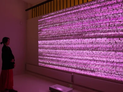 A visitor stands in a dimly lit gallery space facing a monumental digital projection awash in vibrant pink and magenta tones. Layers of overlapping text, handwritten script, and fragmented phrases ripple across the wall-sized display, creating an immersive field of language and light. The installation transforms written words into a dynamic visual landscape, casting a soft glow throughout the room while inviting contemplation of communication, memory, and digital expression. The viewer’s silhouette provides a sense of scale, emphasizing the artwork’s commanding presence and immersive nature