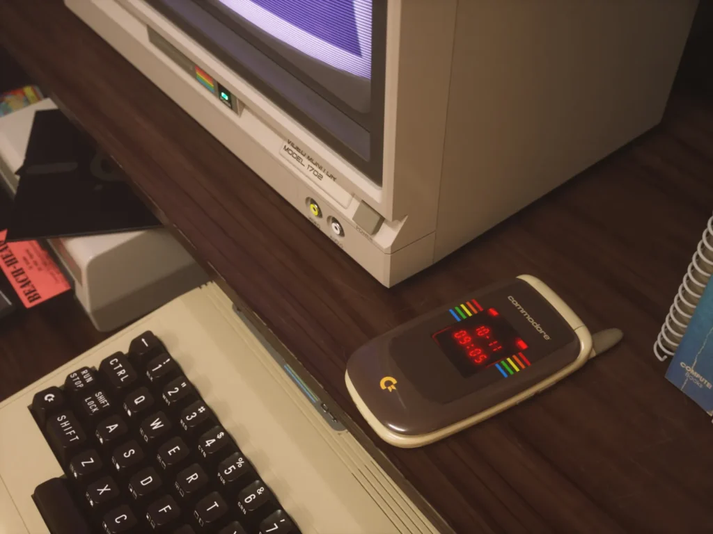 A closed Commodore flip phone sits on a wooden desk beside a vintage Commodore computer, CRT monitor, keyboard, and retro accessories. The phone's external display features red LED graphics and rainbow stripes, reinforcing the connection between classic Commodore computing and the modern concept device