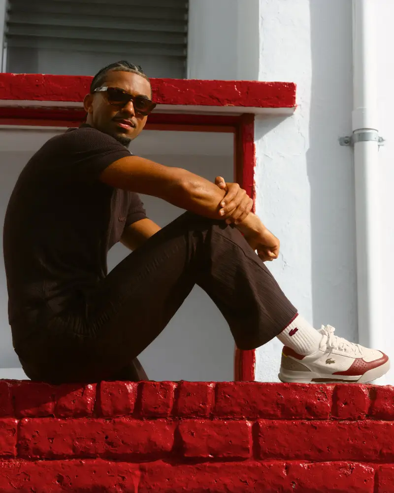 Arthur Fils sits on a bright red brick wall wearing the Lacoste Slam Break Premium sneaker in white and deep red, styled with a dark knit outfit and sunglasses against a sunlit architectural backdrop