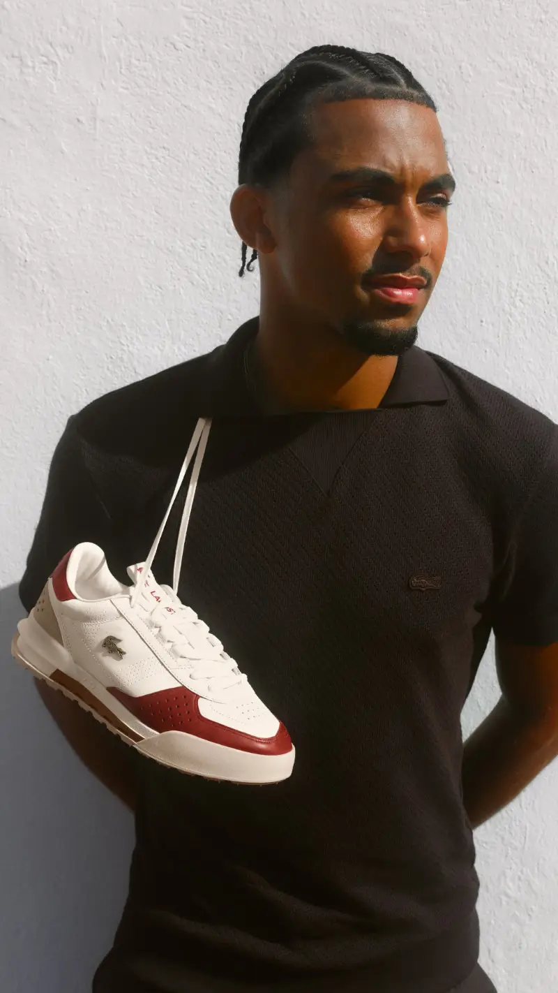 Arthur Fils poses against a white wall while holding the Lacoste Slam Break Premium sneaker, highlighting the shoe's premium leather construction, metallic crocodile logo, and clay-court-inspired red accent