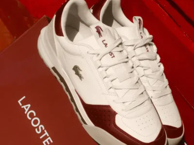 Close-up of the Lacoste Slam Break Premium sneaker displayed beside its premium Lacoste presentation box, showcasing the off-white leather upper, deep red overlays, perforated detailing, and archival crocodile branding
