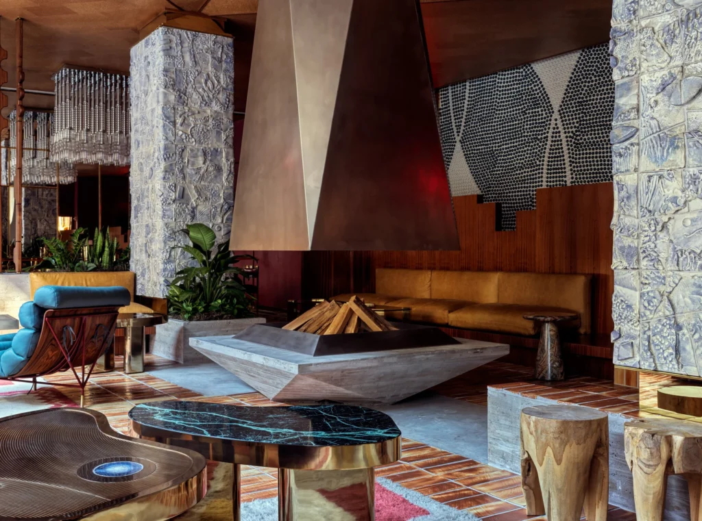 Warmly lit interior lounge with a sculptural central fireplace, textured ceramic columns, rich wood paneling, leather seating, and vintage-inspired furnishing