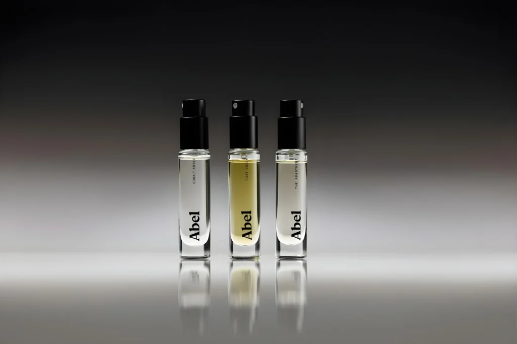 A minimalist product photograph featuring three Abel fragrance travel sprays standing upright on a reflective surface against a dark gradient background. The clear glass vials reveal varying pale-toned liquids and are topped with sleek black atomizer caps, creating a refined, modern luxury aesthetic with subtle reflections and studio light