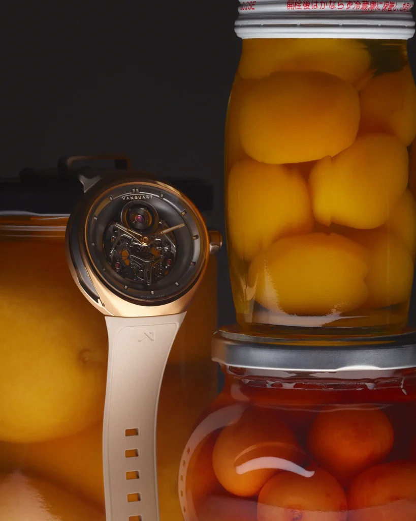 A Vanguart watch with a light-colored strap stands beside stacked jars of preserved fruit. Warm amber lighting highlights the mechanical dial against a backdrop of peaches and cherries suspended in syrup