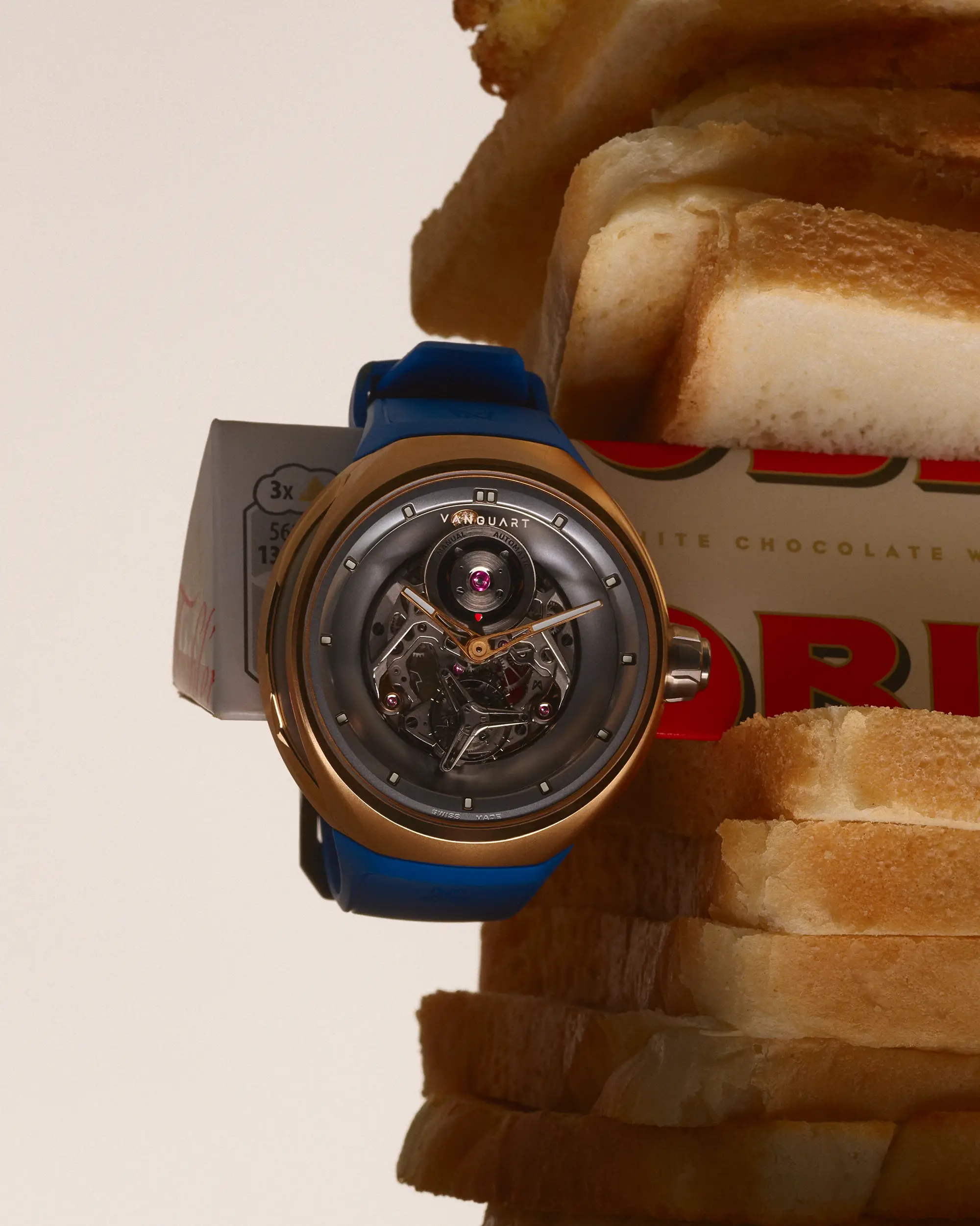 Vanguart watch with a blue strap mounted against stacked slices of white bread and a chocolate bar wrapper. The whimsical arrangement contrasts artisanal watchmaking with familiar pantry staple