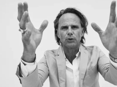 Black-and-white portrait of a man in a light-colored blazer and open-collar shirt facing the camera against a plain background. He extends both hands toward the lens, creating a dramatic sense of depth and motion, while rings and a wristwatch add subtle detail to the minimalist editorial composition