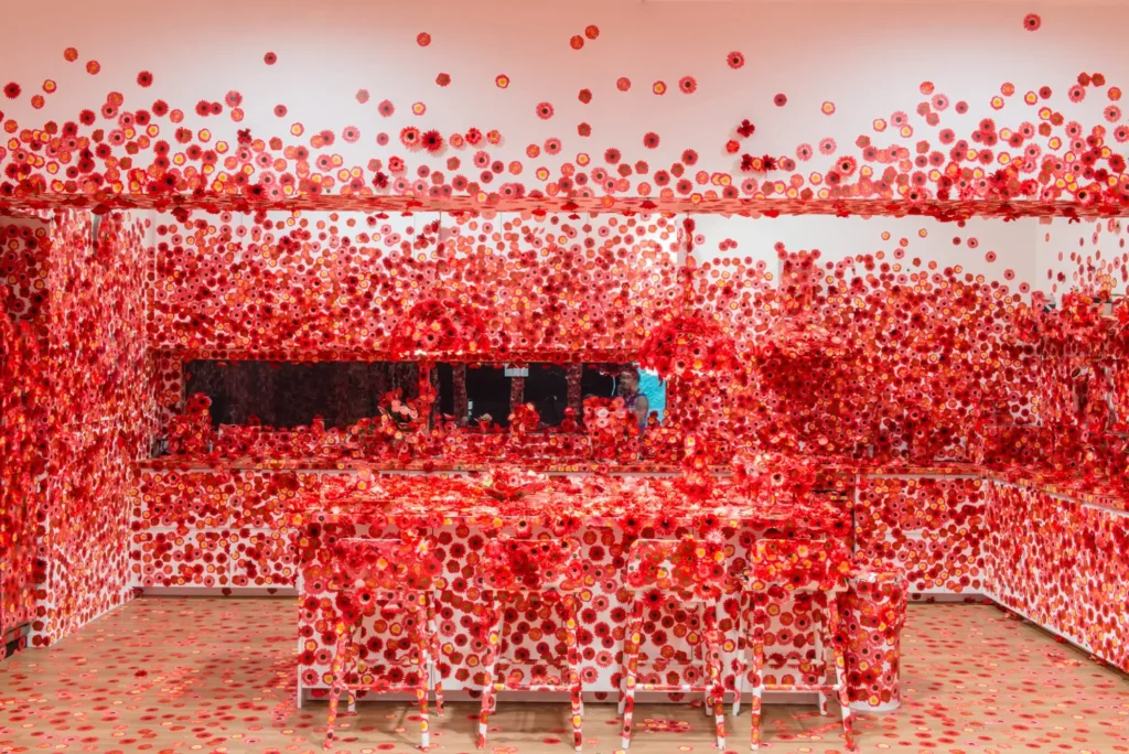 Contemporary art installation by Yayoi Kusama featuring an immersive room covered in thousands of vibrant red floral decals, transforming walls, furniture, ceiling, and decorative objects into a striking polka-dot-inspired environment