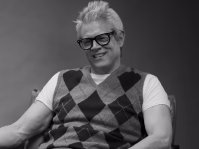 Black-and-white portrait of a smiling Johnny Knoxville seated in a chair during an interview or conversation. They wear thick black rectangular glasses, a white short-sleeve T-shirt, and a gray argyle sweater vest with large diamond patterns. Their light-colored hair is styled upward in a textured quiff. The subject leans back comfortably with one arm resting on the chair, conveying a relaxed, approachable mood against a plain dark studio background
