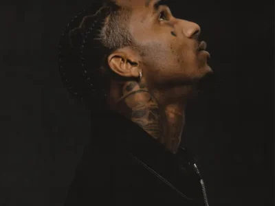 Side-profile portrait of rapper Loe Shimmy against a dark background, wearing a black jacket and looking upward. His braided hairstyle, facial tattoos, neck tattoo, and earring are visible, creating a moody and introspective promotional image