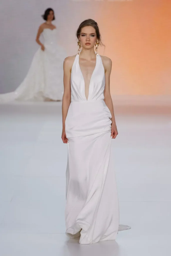 Bridal runway look featuring a sleek white halter-neck gown with a dramatic plunging neckline, softly draped skirt, and statement gold earring