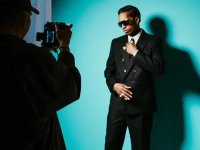Fashion photoshoot featuring A$AP Rocky posing in a tailored black double-breasted suit, white shirt, and tie against a vibrant teal backdrop. Wearing dark sunglasses and jewelry, he adjusts his lapel while a photographer captures the scene, casting a dramatic shadow that enhances the sleek, editorial atmosphere