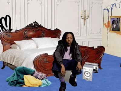 Alt text: A musician sits in front of an ornate vintage wooden bed in a stylized bedroom set featuring white paneled walls, blue carpet, scattered clothing, a keyboard on a stand, and retro audio equipment. Graffiti-style artwork and framed childhood photos decorate the wall, creating a personal, nostalgic atmosphere that blends luxury furnishings with creative studio element
