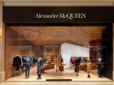 Alexander McQueen luxury boutique storefront featuring floor-to-ceiling glass windows, warm wood interiors, tailored menswear and womenswear displays, and the brand’s illuminated logo above the entrance