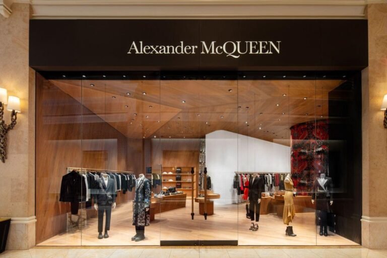 Alexander McQueen luxury boutique storefront featuring floor-to-ceiling glass windows, warm wood interiors, tailored menswear and womenswear displays, and the brand’s illuminated logo above the entrance