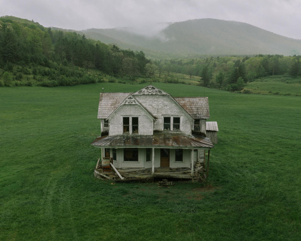 A deteriorating white farmhouse stands alone in a lush green meadow, backed by mist-covered mountains and rolling countryside under a rainy sky