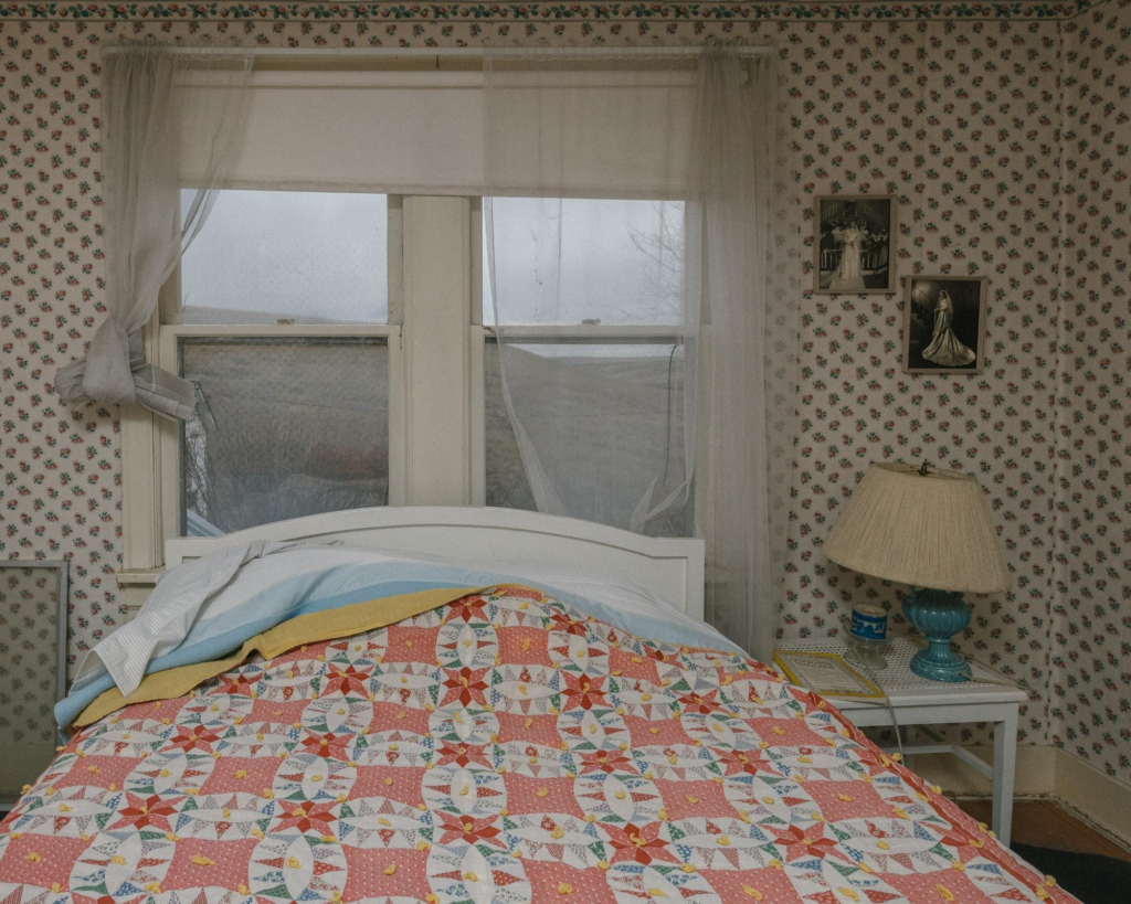 A neatly made bed with a colorful patchwork quilt sits in a vintage bedroom, framed by floral wallpaper, sheer curtains, and soft natural light from the window