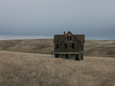 A weathered abandoned farmhouse stands isolated in tall prairie grass, surrounded by gently rolling hills under a heavy gray sky