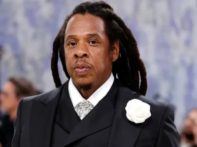 Portrait of a man with long dreadlocks wearing a tailored black double-breasted suit, white shirt, ornate silver neckpiece, and large white floral lapel pin. He faces the camera with a composed expression while standing in front of a softly blurred event backdrop, creating an elegant red-carpet-style image