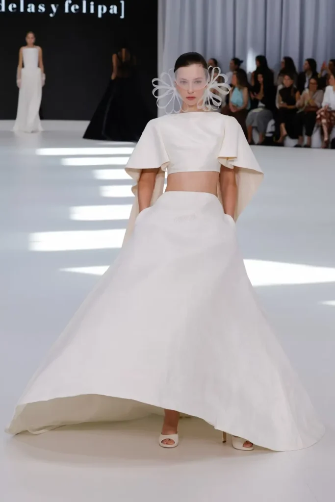 Modern bridal ensemble with a cropped cape-sleeve top, voluminous ball skirt, and sculptural floral-inspired face accessory showcased on the runway
