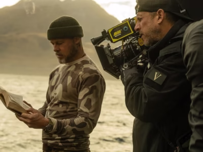 A behind-the-scenes production still shows an actor standing by a lake with mountains in the background, reading from an open book while wearing an olive beanie, camouflage long-sleeve shirt, and wristwatch. Beside him, a cinematographer films the scene using an ARRI cinema camera, wearing a black jacket and backward cap. The golden-hour lighting and rugged landscape create a cinematic atmosphere during the outdoor shoot