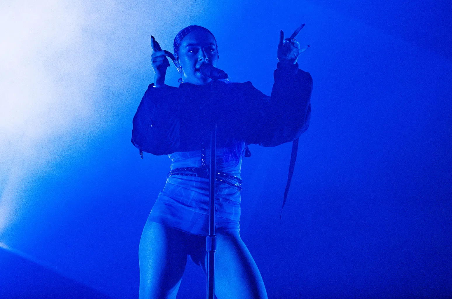 A performer sings into a microphone on a dimly lit stage bathed in intense blue lighting and atmospheric haze. Wearing an oversized dark top and a fitted light-colored outfit, the artist gestures toward the crowd while standing at a microphone stand, creating a dramatic concert scene