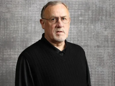 Portrait of legendary NBA coach Rick Adelman wearing glasses and a black collared shirt layered with a textured black vest, standing against a gray patterned backdrop. The Hall of Fame coach looks directly toward the camera with a calm and thoughtful expression, reflecting the leadership and basketball intelligence that defined his decades-long career with the Portland Trail Blazers, Sacramento Kings, Houston Rockets, and Minnesota Timberwolves