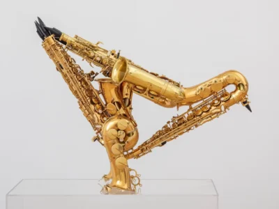 Three polished brass saxophones arranged in an interlocking triangular sculpture on a clear acrylic pedestal. The instruments feature reflective gold finishes, intricate keywork, black mouthpieces, and overlapping bells against a clean white background, creating a striking display that blends musical craftsmanship with contemporary sculptural design