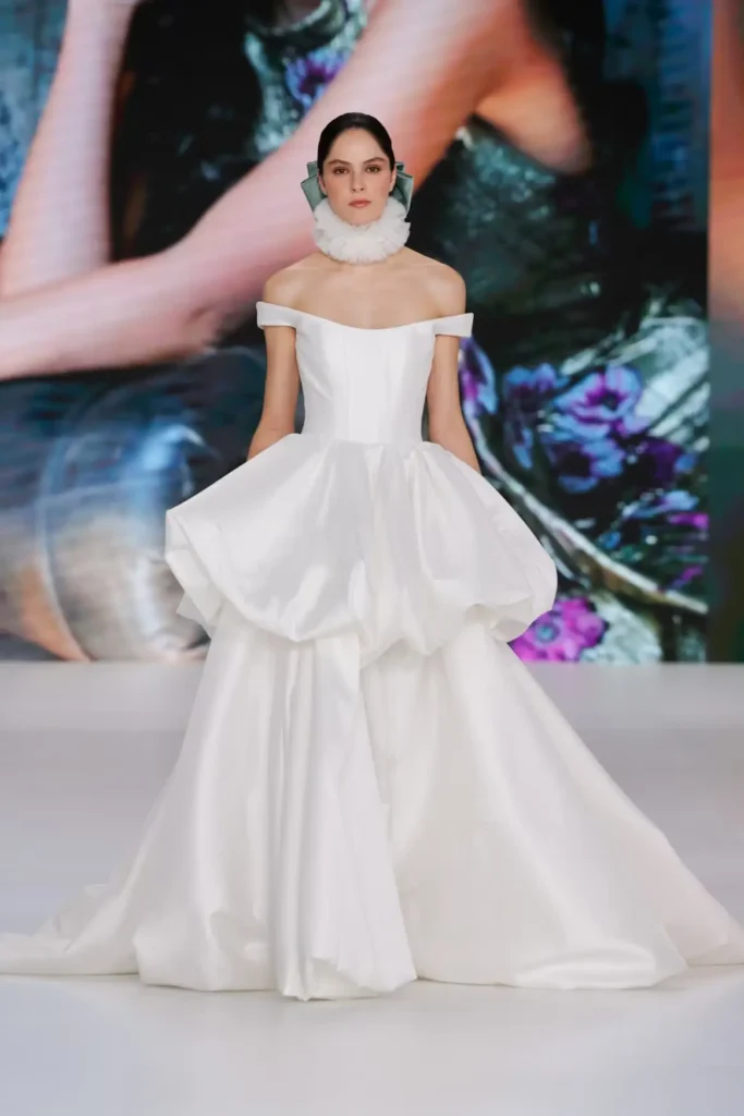 Off-the-shoulder bridal ball gown with dramatic puffed overskirt, flowing train, and ruffled neck accessory on the runway