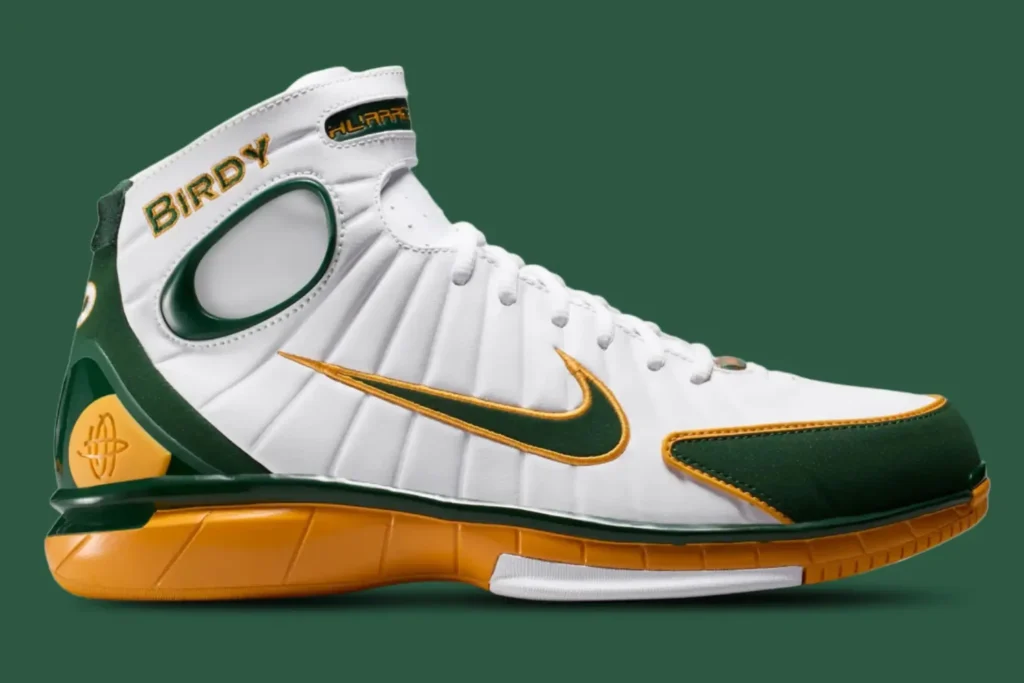 Side profile of the Nike Zoom Huarache 2K4 “Birdy” basketball shoe in white leather with green and gold accents, featuring Larry Bird-inspired branding, embroidered Swoosh detailing, and a golden performance midsole against a green background
