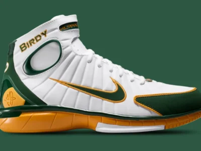 Side profile of the Nike Zoom Huarache 2K4 “Birdy” basketball shoe in white leather with green and gold accents, featuring Larry Bird-inspired branding, embroidered Swoosh detailing, and a golden performance midsole against a green background