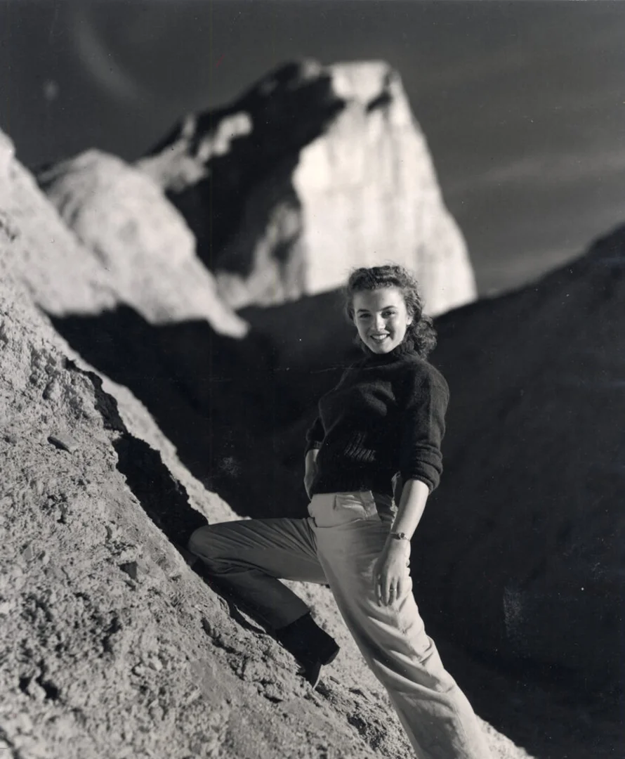 Black-and-white photograph of a young Marilyn Monroe posing on a rugged mountainside, wearing high-waisted trousers and a dark sweater, with dramatic rock formations rising behind her in a striking outdoor portrait