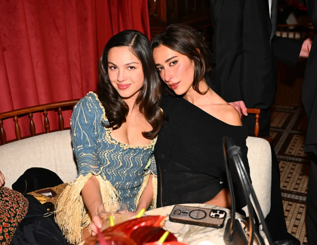 Two women sit closely together on a cream-colored sofa at an intimate evening gathering. One wears a textured blue dress with fringe detailing, while the other wears a black off-the-shoulder knit top. Rich red drapery and warm lighting create an elegant, upscale atmosphere around the table set