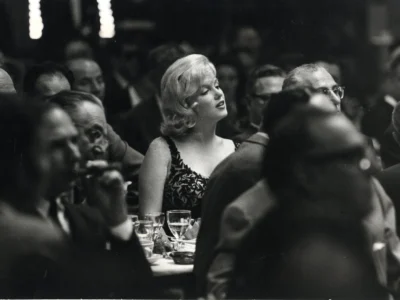 Black-and-white photograph of Marilyn Monroe seated among a crowd at a formal dinner event, her light-colored hair and elegant patterned evening dress drawing focus amid the dimly lit room filled with guests in suits and gowns