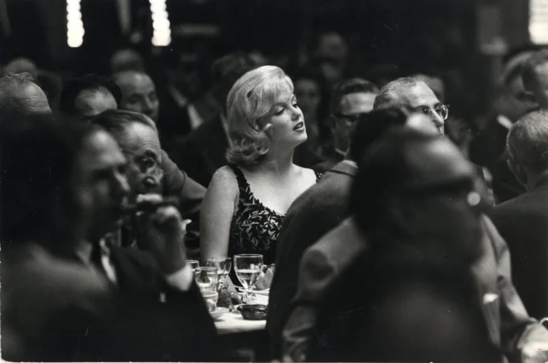 Black-and-white photograph of Marilyn Monroe seated among a crowd at a formal dinner event, her light-colored hair and elegant patterned evening dress drawing focus amid the dimly lit room filled with guests in suits and gowns