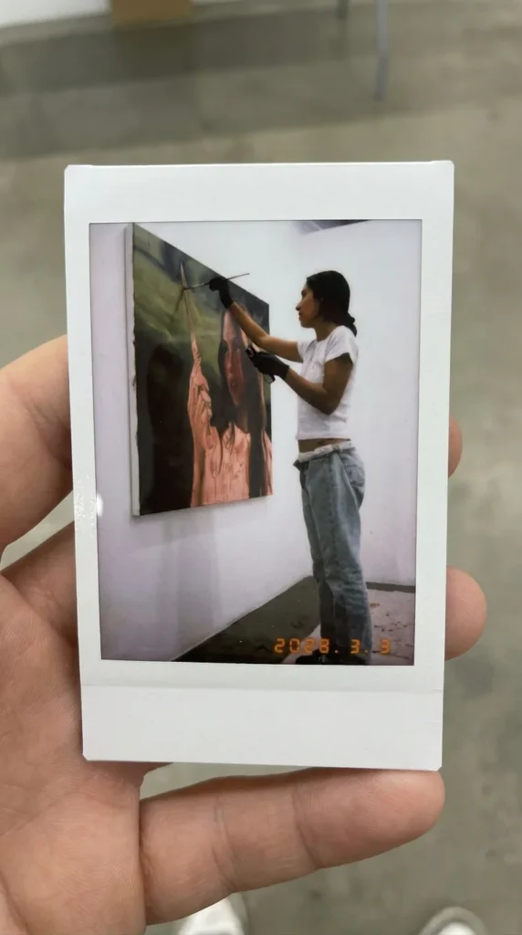 A hand holds a Polaroid-style instant photograph showing an artist working on a large portrait painting in a contemporary gallery space. The artist stands beside the canvas wearing casual clothing and gloves, captured mid-process in a bright, minimalist exhibition set