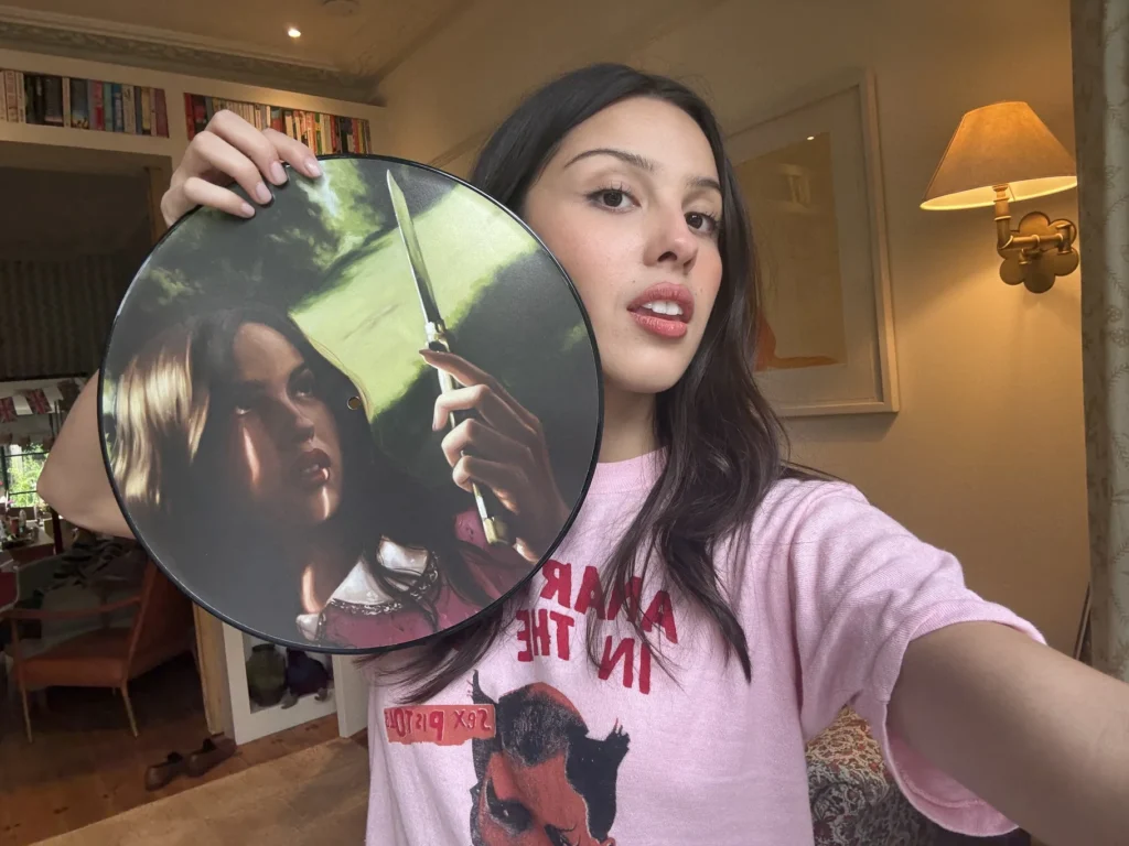 A woman takes a selfie indoors while holding a circular artwork featuring a dramatic portrait painted in rich, cinematic tones. Warm ambient lighting, bookshelves, and framed wall art create a cozy home environment in the background