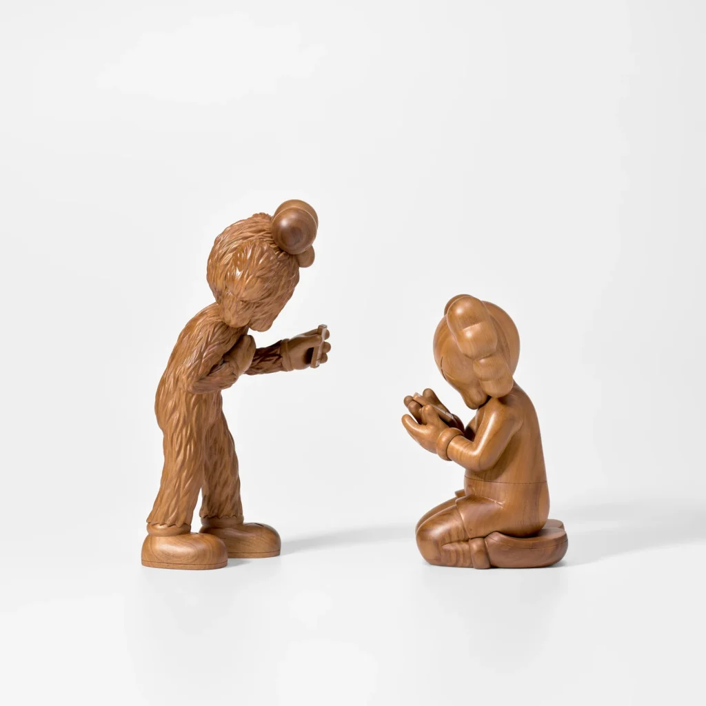 Polished wooden sculptures inspired by iconic pop-culture characters face one another in a contemplative scene, with one standing and holding a small object while the other kneels, both crafted from richly grained natural wood against a minimalist gallery-style backdrop
