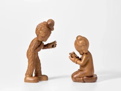 Polished wooden sculptures inspired by iconic pop-culture characters face one another in a contemplative scene, with one standing and holding a small object while the other kneels, both crafted from richly grained natural wood against a minimalist gallery-style backdrop