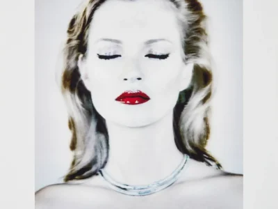 A high-key fashion portrait of a blonde model with her eyes closed, wearing vivid glossy red lipstick and a minimalist silver necklace. The image features soft, luminous lighting that nearly washes out the skin tones, creating an ethereal editorial aesthetic. Her hair is swept back in loose waves, while bold winged eyeliner and the vibrant red lips provide striking contrast against the pale monochrome palette. The portrait is centered within a wide white border and appears as a fine art print, with a subtle artist signature visible in the lower-right corner. The composition emphasizes elegance, serenity, and classic beauty with a modern, gallery-style presentation