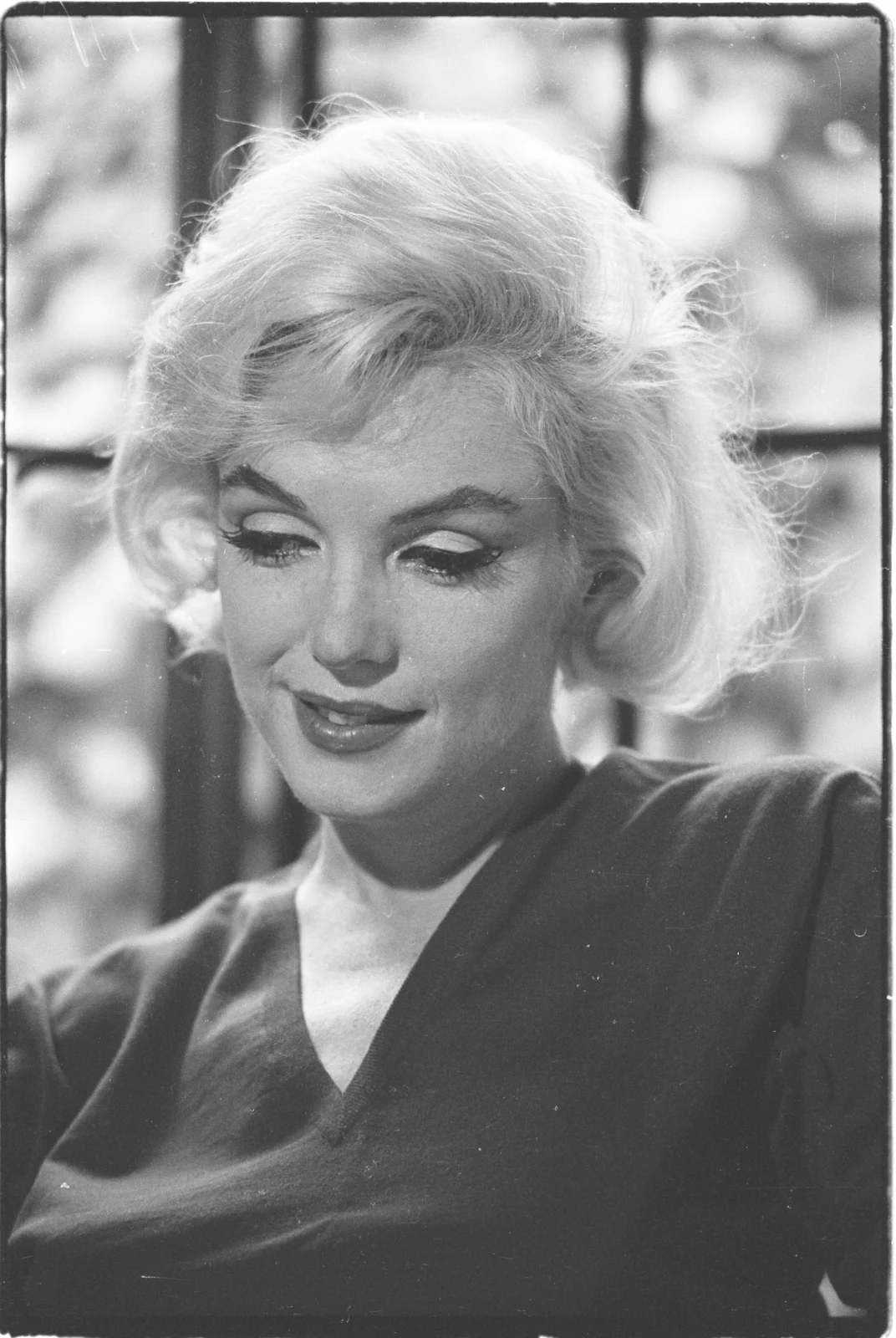 Close-up black-and-white portrait of Marilyn Monroe with softly styled platinum hair, dramatic eye makeup, and a contemplative expression, illuminated by natural light from a nearby window