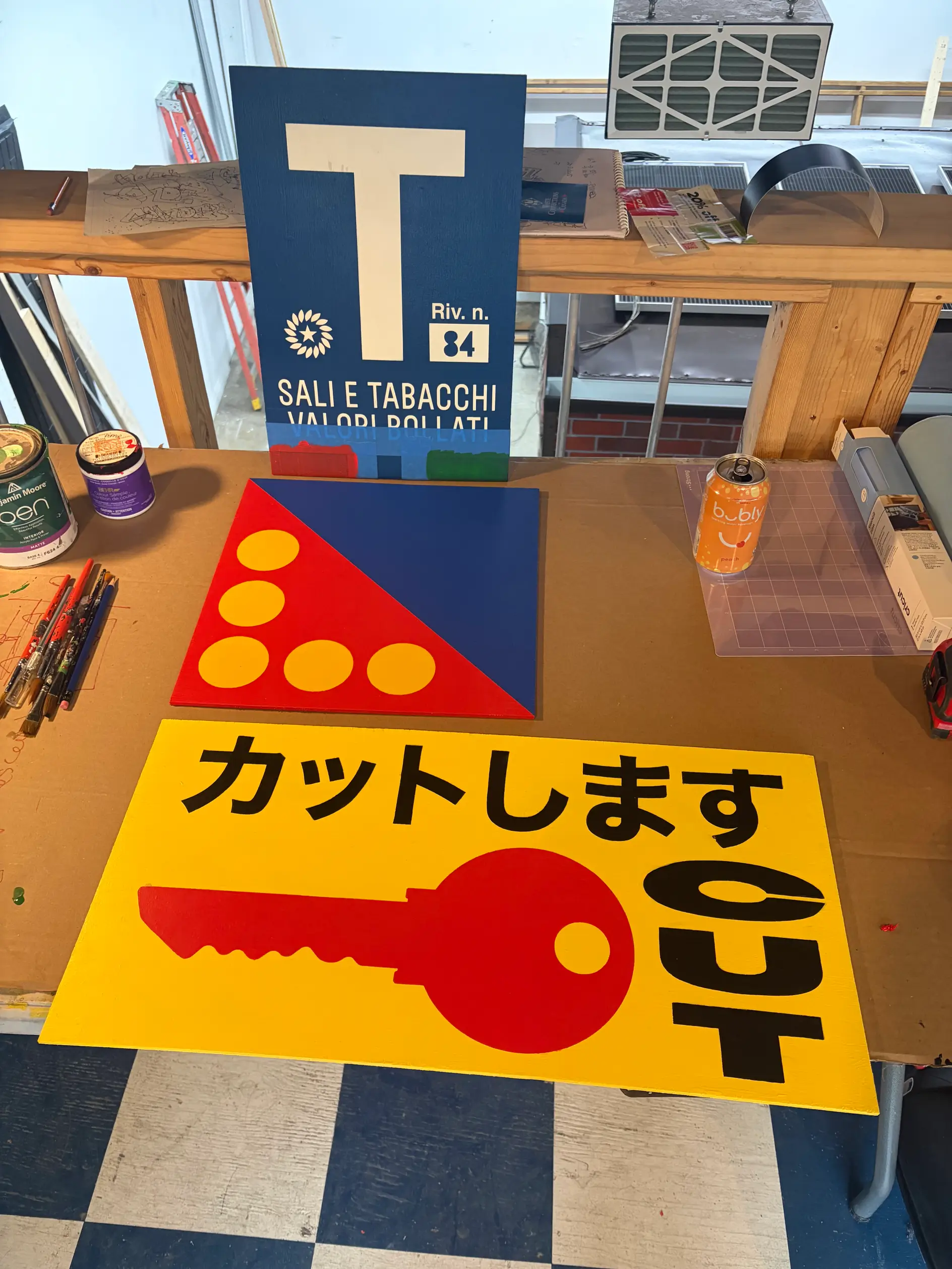 Custom-made graphic signs and painted panels displayed on a workshop table, including multilingual convenience-store-inspired artwork used throughout the install