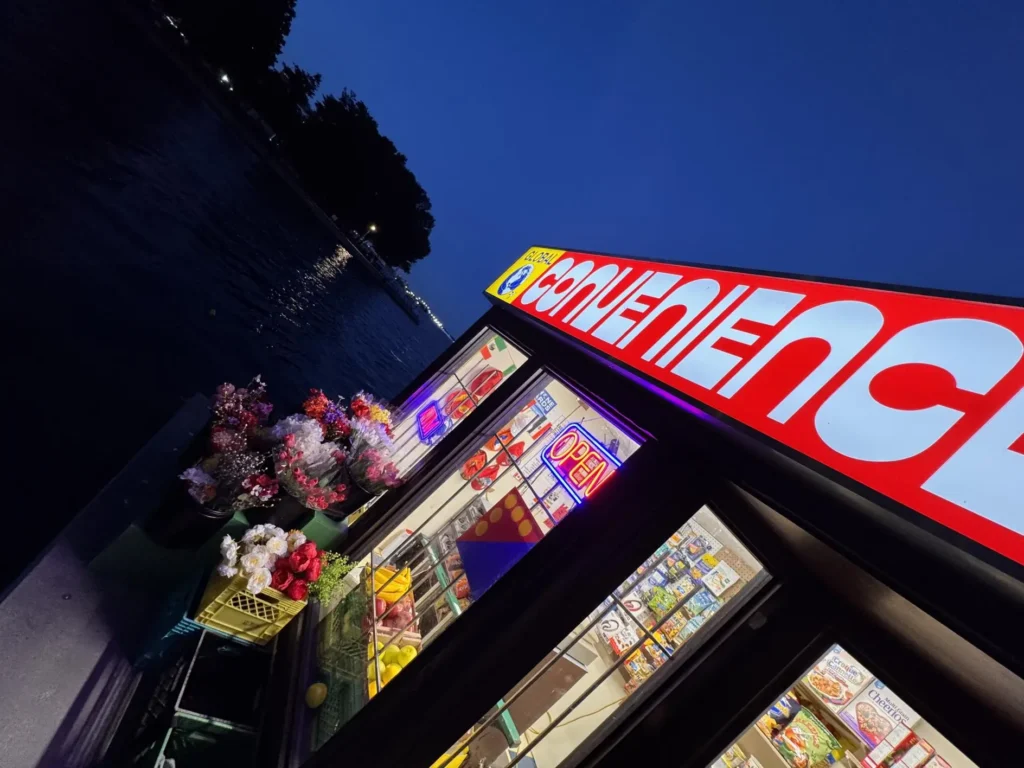 Dramatic low-angle nighttime view of the floating convenience store, highlighting the glowing sign, neon lights, storefront windows, and surrounding dark waterfront atmosphere
