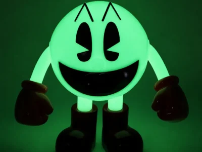Glow-in-the-dark PAC-MAN collectible figure illuminated in bright green, standing against a dark background. The character’s classic smiling face, black eyes, red gloves, and oversized boots create a nostalgic arcade-inspired display with a vivid neon glow effect