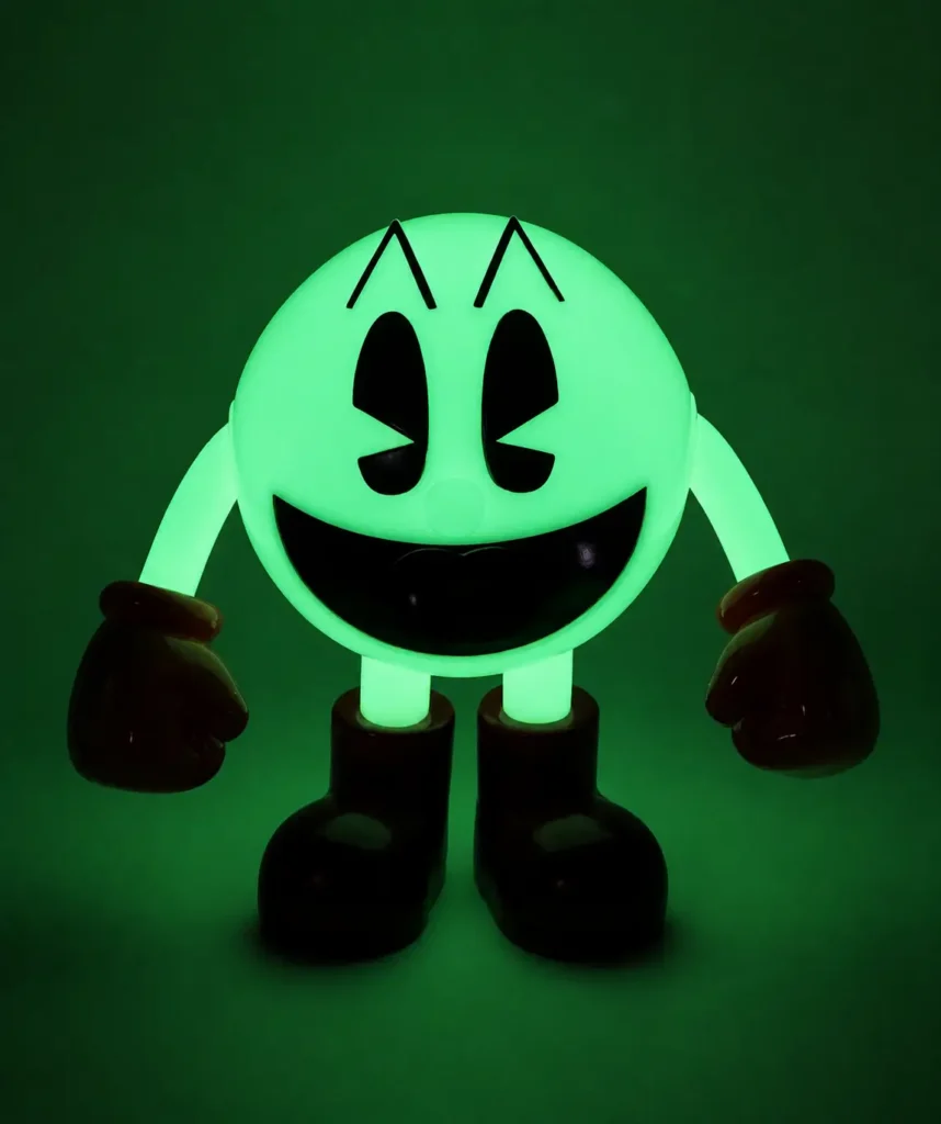 Glow-in-the-dark PAC-MAN collectible figure illuminated in bright green, standing against a dark background. The character’s classic smiling face, black eyes, red gloves, and oversized boots create a nostalgic arcade-inspired display with a vivid neon glow effect