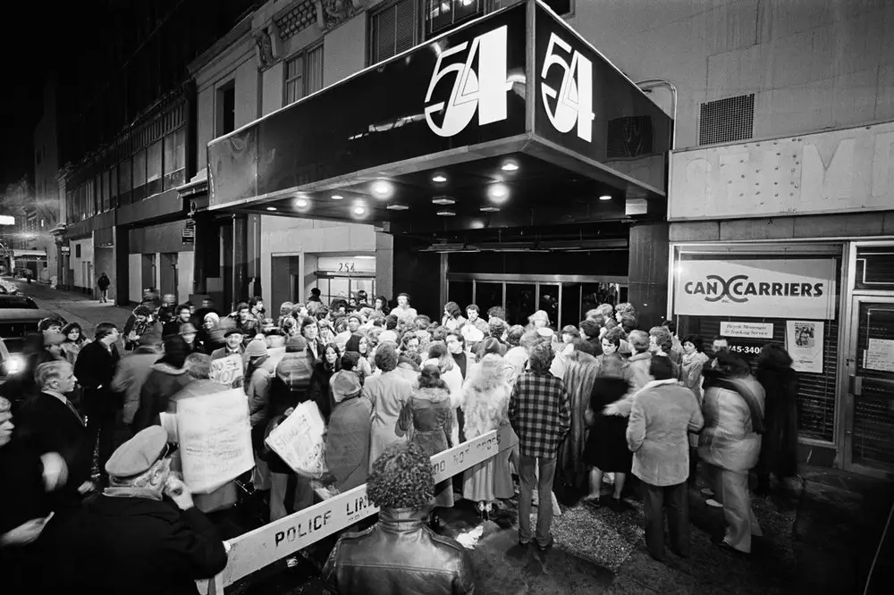 Historic black-and-white photograph of the exterior of Studio 54 in New York City, showing a large crowd gathered outside the iconic nightclub beneath its illuminated marquee. Police barricades line the entrance as attendees wait on the sidewalk, capturing the excitement, exclusivity, and cultural significance of one of the most famous nightlife venues of the 1970s