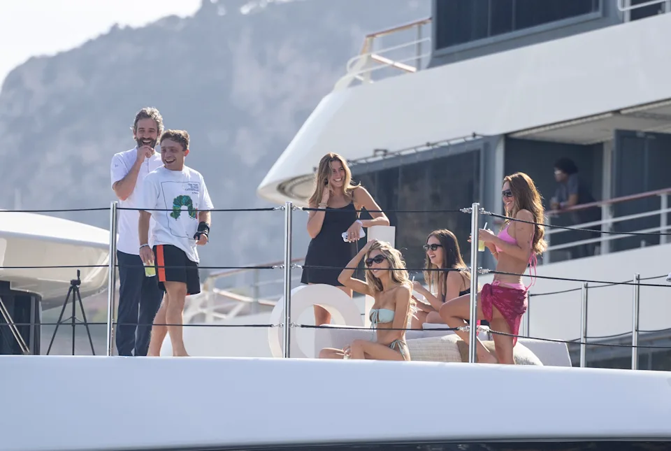 A group of influencers and guests relax aboard a luxury yacht in the Mediterranean, chatting and enjoying the sunshine. Swimwear, casual resort attire, and scenic coastal views create an atmosphere of exclusive summer leisure