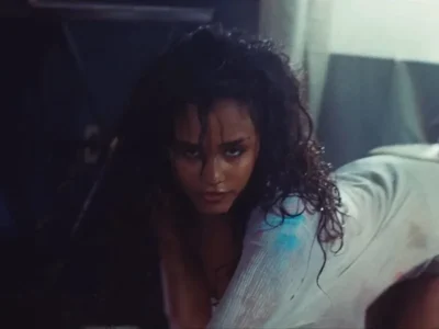 Moody still from Tyla’s “IS IT LOVE” music video showing the singer with wet, tousled hair and an intense gaze while crouched on the floor in a sheer white outfit. Dim blue-toned lighting, reflective surfaces, and cinematic sil create an atmosphere of vulnerability, desire, and emotional tension