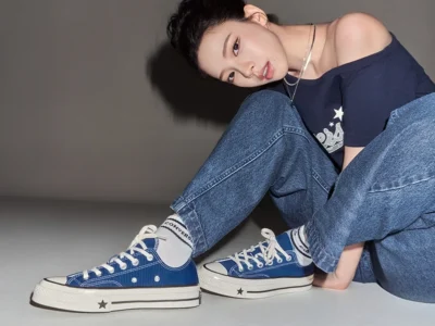 A model lounges on the floor wearing a black Converse graphic T-shirt, flowing white skirt, gray scrunched socks, and black Converse Chuck Taylor high-top sneakers with silver chain accessories. The minimalist studio setting and relaxed pose highlight the casual streetwear styling and iconic canvas footwear