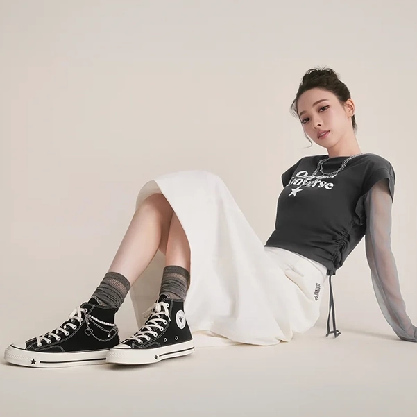 Karina of aespa reclining in a minimalist studio wearing a black Converse graphic T-shirt, white midi skirt, gray scrunched socks, and black Converse Chuck Taylor All Star high-top sneakers accented with silver chain accessories.