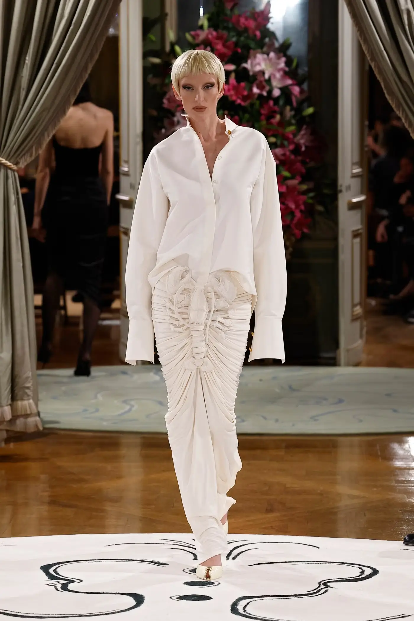 Runway model walking through an ornate salon wearing an oversized white shirt paired with a sculptural white skirt shaped like a lobster, combining tailoring and surrealist design